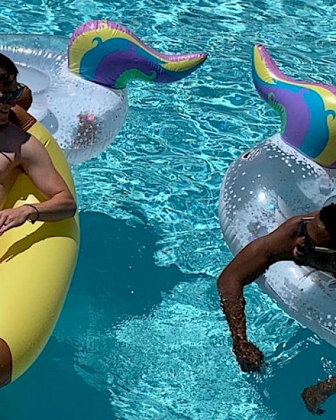 Two men are floating in a pool; one on a yellow float and the other on a unicorn float. Both are enjoying a sunny day.